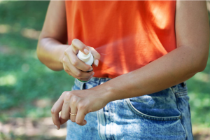 A woman applies insect repellent spray to her arm in a woodland area.