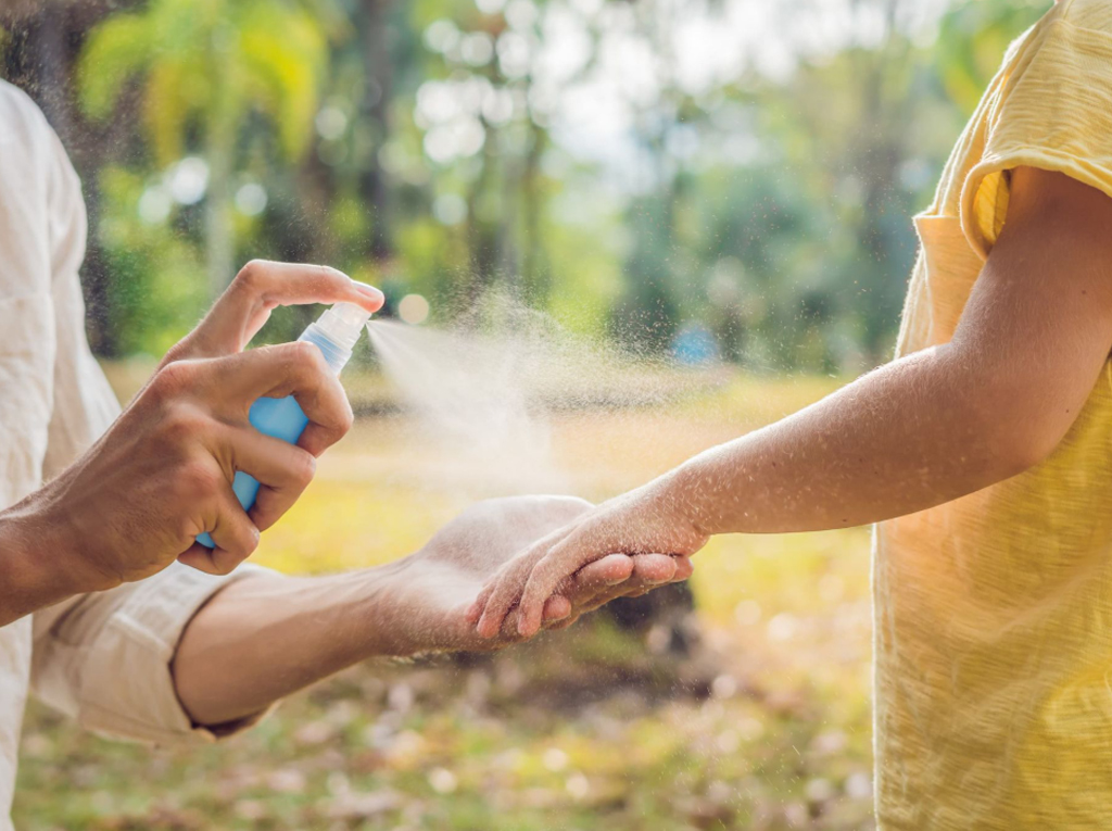 A man sprays insect repellent onto his child’s arm in a forest clearing.