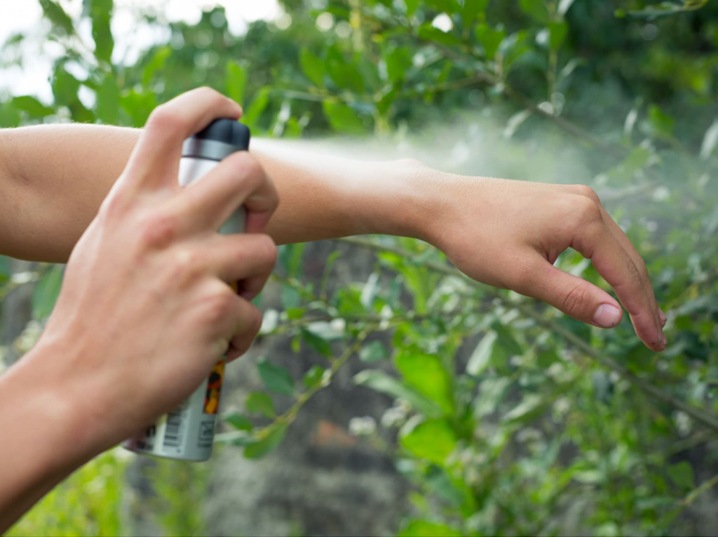 A close-up of someone applying an insect repellent spray to their wrist in a lush, green outdoor environment.